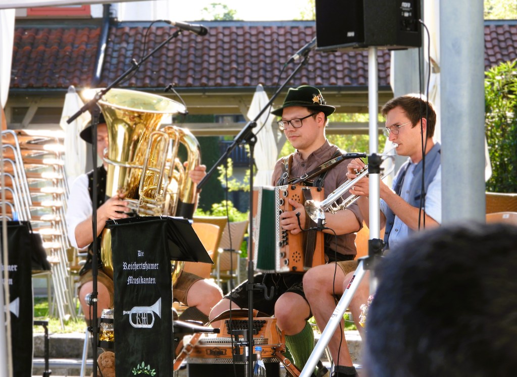 Die Reichertshamer Musikanten spielen auf der Bühne, mit einem Tuba-Spieler, der in traditioneller bayerischer Tracht gekleidet ist, und weiteren Musikern, die verschiedene Instrumente spielen.