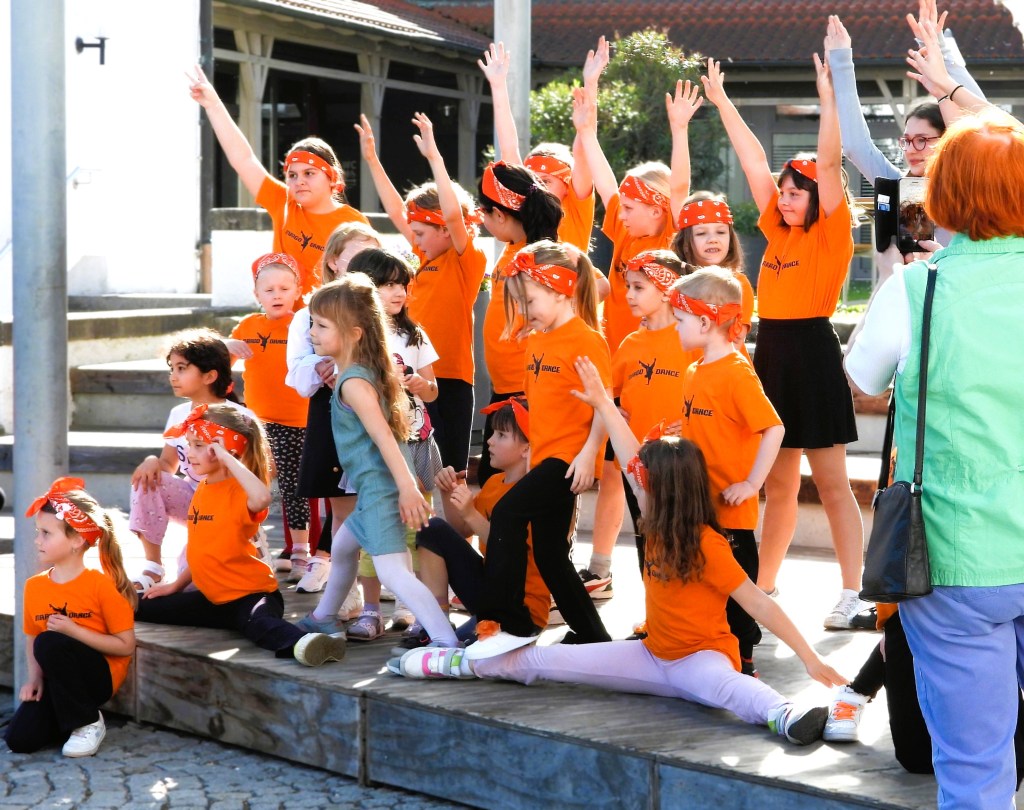Eine Gruppe von Kindern in orangefarbenen T-Shirts und Kopftüchern steht auf einer Holzplattform. Einige Kinder haben die Arme erhoben und posieren fröhlich, während andere auf dem Boden sitzen oder liegen.