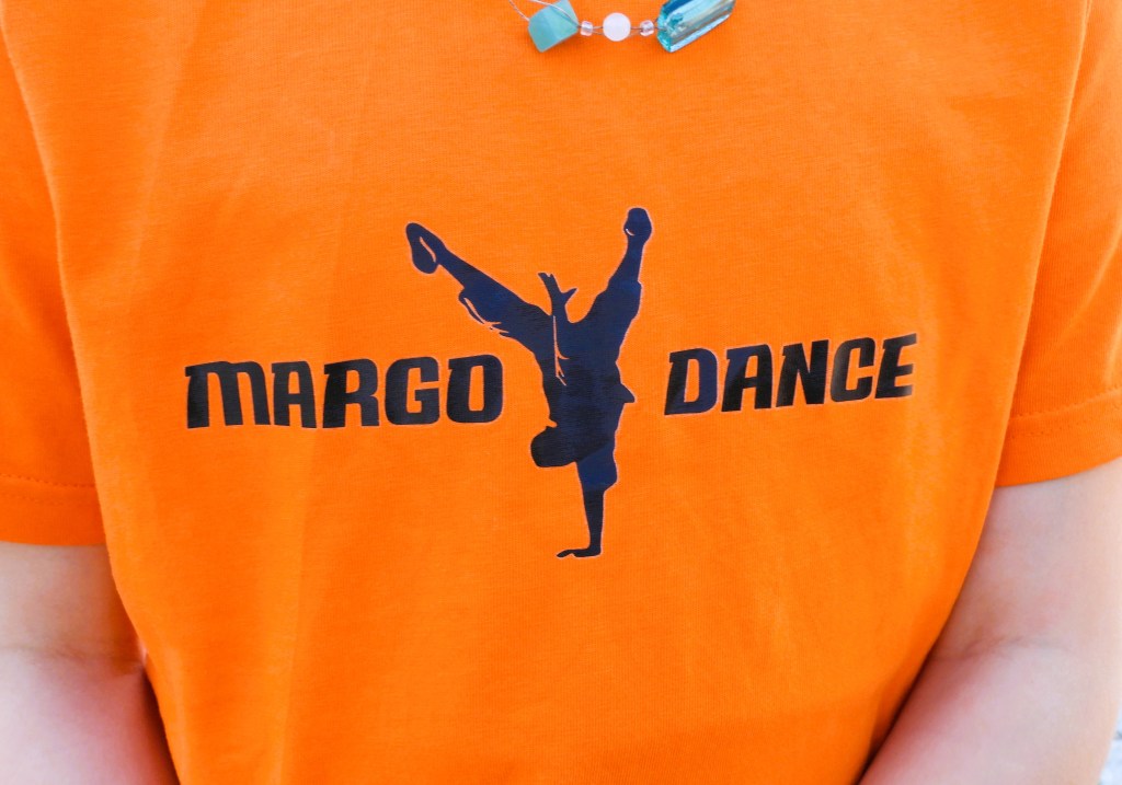 Nahaufnahme eines orangefarbenen T-Shirts mit dem Aufdruck 'MARGO DANCE' und einer tanzenden Silhouette.