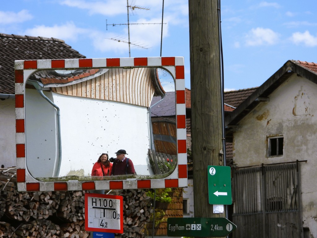 Spiegel mit reflektierenden Bildern von zwei Personen, die in einer ländlichen Umgebung stehen, umgeben von Holzstapeln und alten Gebäuden.