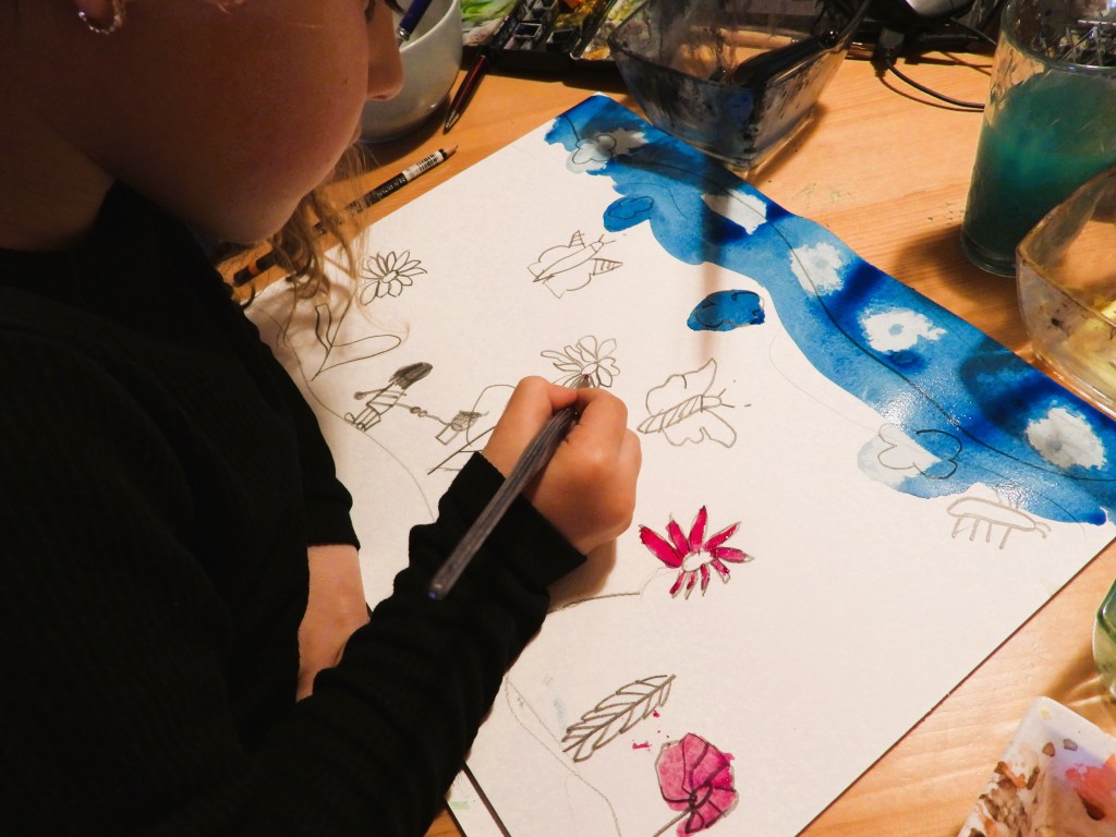 Ein Kind malt mit einem Pinsel auf einem Blatt Papier, während um sie herum Malutensilien und Farben liegen.