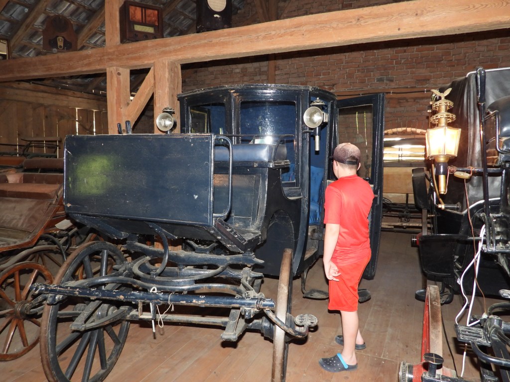 Ein Junge in rotem Shirt und Shorts steht vor einer alten Kutsche in einem Bauernhofmuseum.