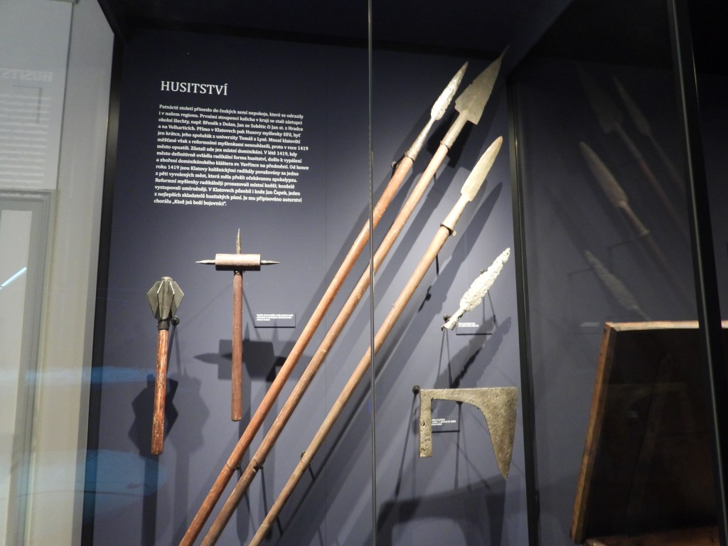 Historische Waffen und Werkzeugexponate aus der Hussitenzeit in einem Museum, darunter Speere, eine Streitaxt und andere Werkzeuge, ausgestellt auf einem dunklen Hintergrund mit einer erklärenden Tafel.