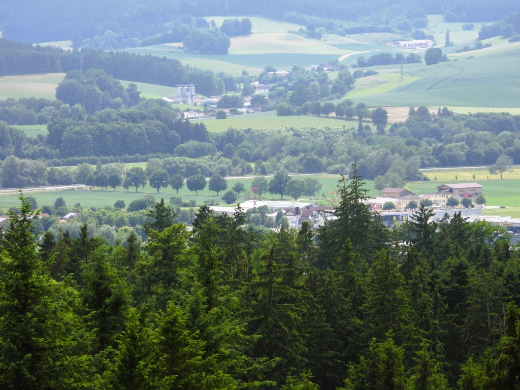 A scenic view of a lush green valley surrounded by rolling hills and forests, with a small village nestled at the foot of the trees under bright, sunny skies.