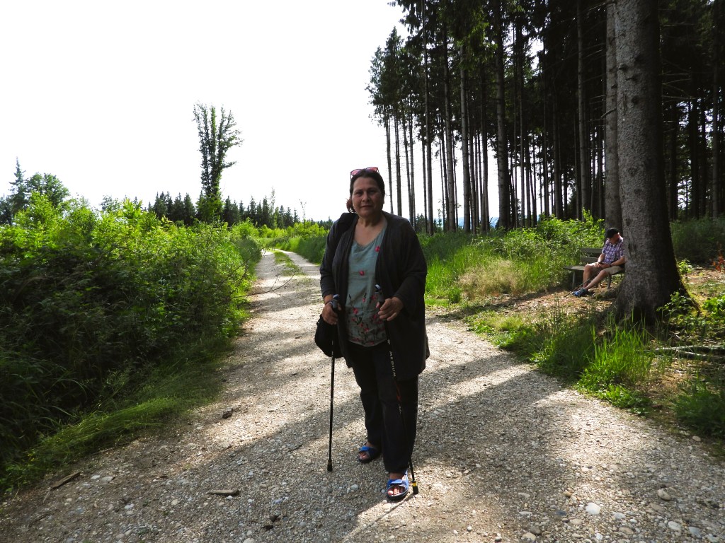 Eine Frau steht auf einem Wanderweg im Wald, umgeben von hohen Bäumen und üppigem Grün, während ein Mann im Hintergrund auf einer Bank sitzt.