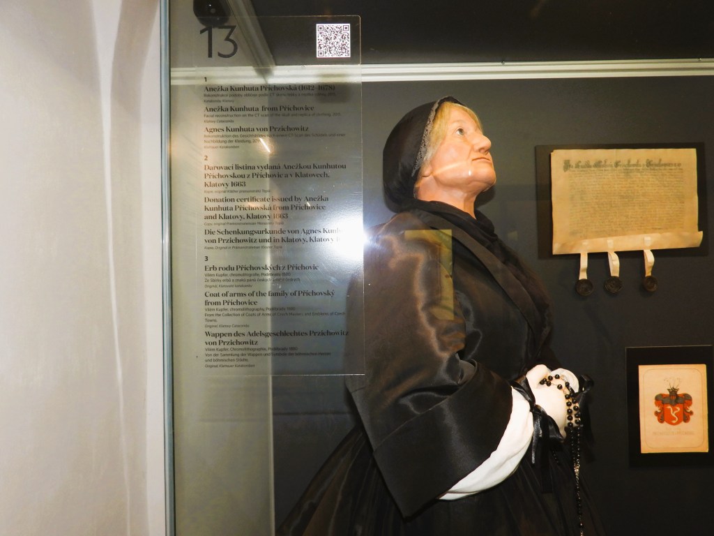 Eine Wachsfigur von Agnes Kunhuta Przichowska, gekleidet in einem schwarzen Kleid, steht neben einem Dokument und einem Wappen in einem Museum.