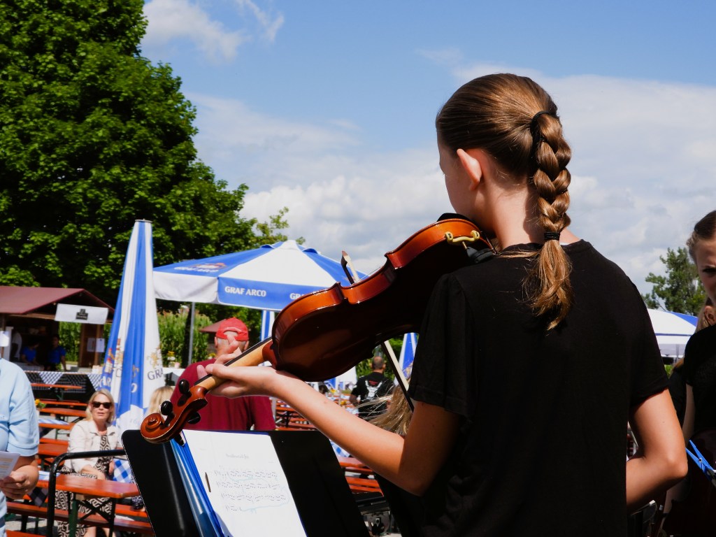 Ein Mädchen mit Zopf hält eine Geige auf einem Sommerfest, während im Hintergrund andere Menschen und Bäume zu sehen sind.