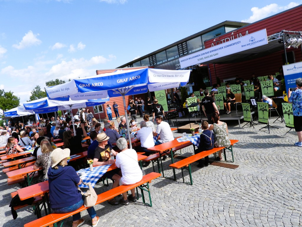 Menschen sitzen an Tischen unter Sonnenschirmen beim Bad Birnbacher Sommerfest, auf einer Bühne treten Musiker auf.
