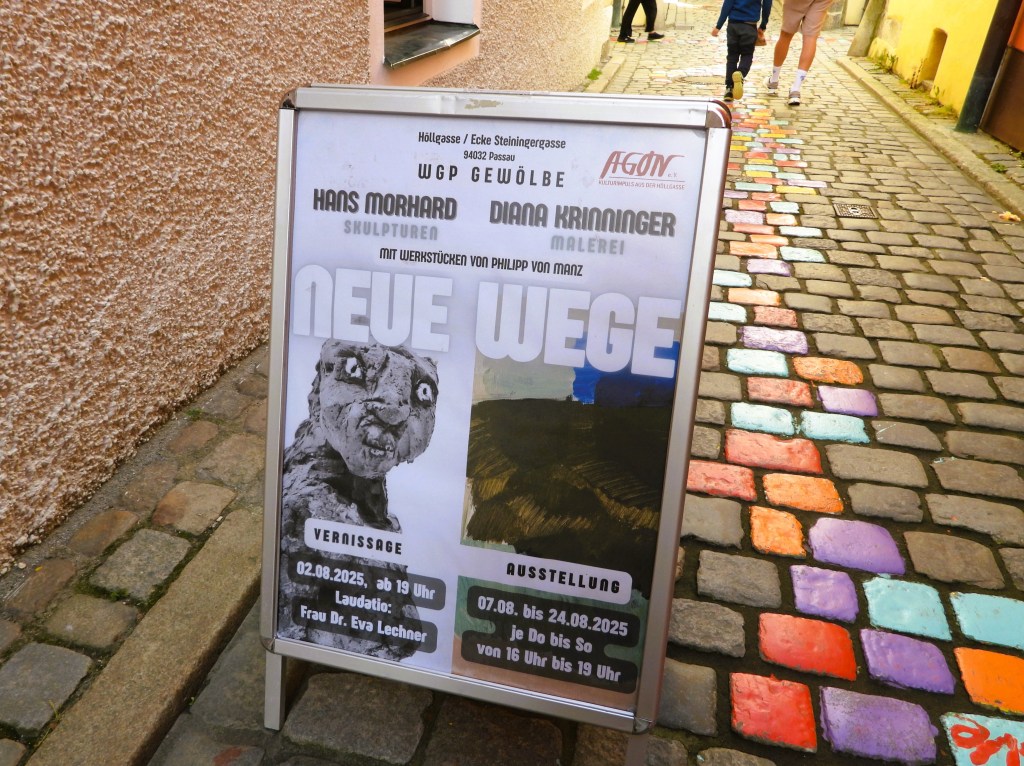 Ein Werbeschild für eine Kunstausstellung in Passau, mit den Namen der Künstler Hans Morhard und Diana Krinninger, im Hintergrund sind bunten Pflastersteine zu sehen.