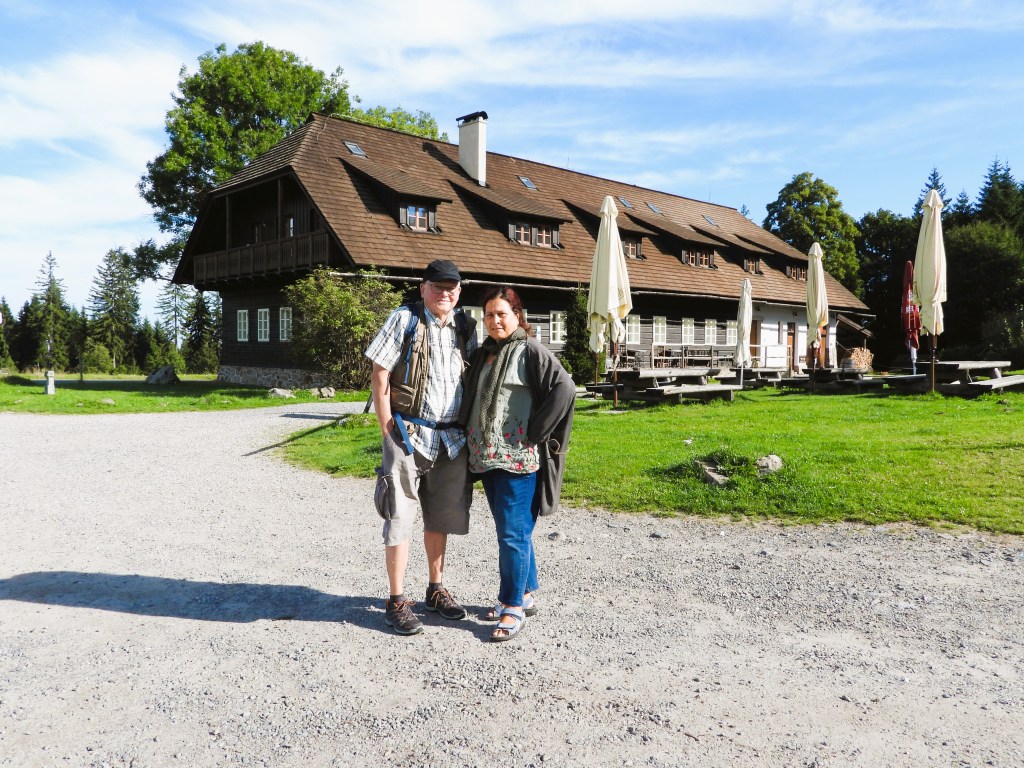 Ein Paar steht lächelnd vor einem großen, traditionell gestalteten Haus im Böhmerwald. Das Gebäude hat ein schräges Dach und ist von einem grünen Rasen umgeben. Im Hintergrund sind Bäume und einige Tischdecken auf den Tischen sichtbar.