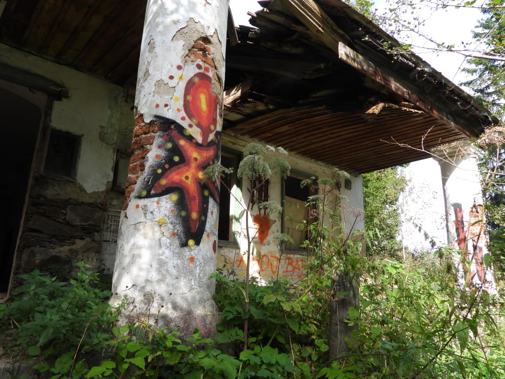 Verfallenes Gebäude im Böhmerwald mit farbenfrohen Graffiti und umgebender Wildvegetation.