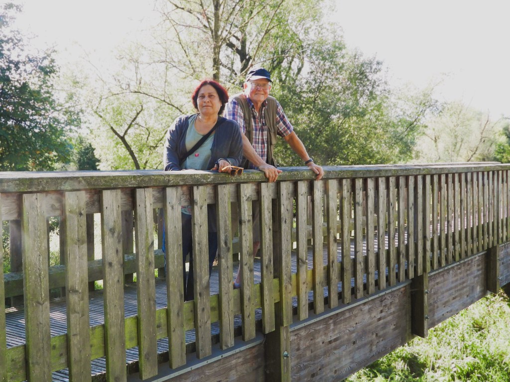 Zwei Personen stehen auf einer Holzbrücke, umgeben von grünen Bäumen und einem klaren Himmel, und schauen in die Ferne.