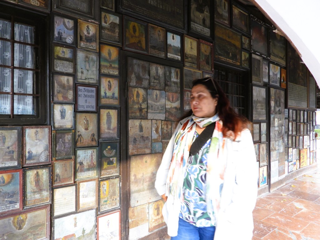 Eine Person steht an einer Wand, die mit zahlreichen Votivbildern dekoriert ist, darunter Geschichten von Dankbarkeit und Heilung.