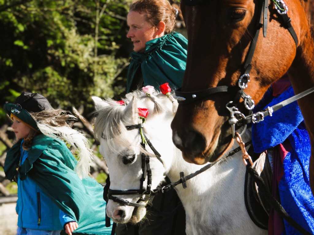 Eine Frau und ein Kind begleiten geschmückte Pferde während der Leonhardiritt-Prozession, umgeben von grüner Natur.