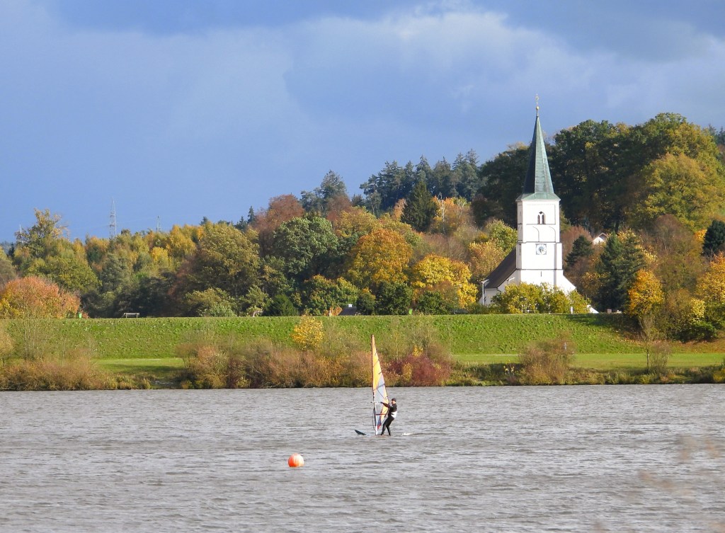 Ein Windsurfer kämpft gegen den Wind auf einem ruhigen See, mit buntem Laub im Hintergrund und einer Kirche im Hintergrund, während der Himmel dramatische Wolken zeigt.