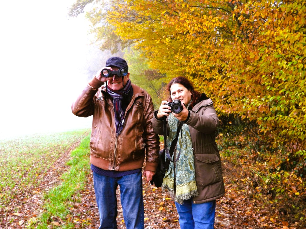 Ein älteres Paar steht auf einem herbstlichen Weg, der von buntem Laub gesäumt ist, während die Frau mit einer Kamera und der Mann mit einem Fernglas in der Hand lächelt.