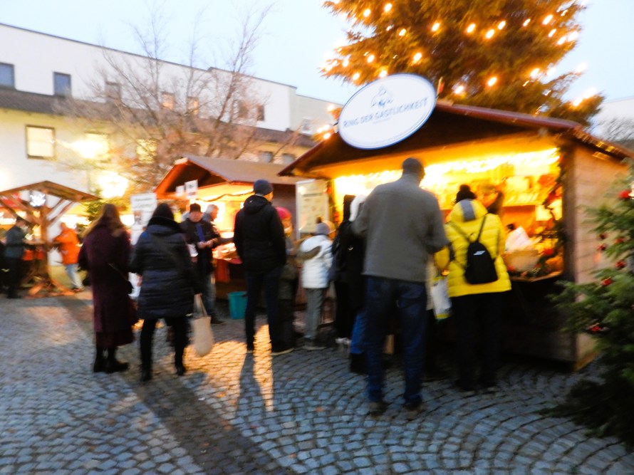 Weihnachtsmarkt mit mehreren Holzständen, die festlich beleuchtet sind, und einer Menschenmenge vor den Ständen.