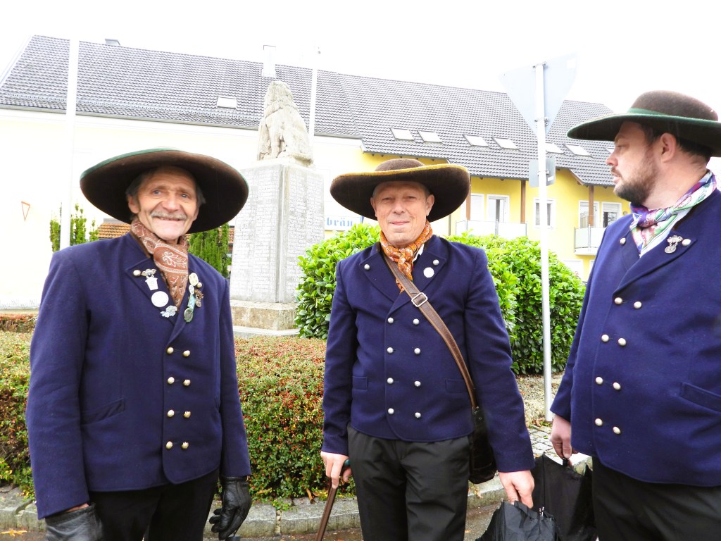 Drei Männer in traditioneller bayerischer Tracht mit großen Hüten stehen vor einer Statue, umgeben von grünen Sträuchern und Gebäuden.
