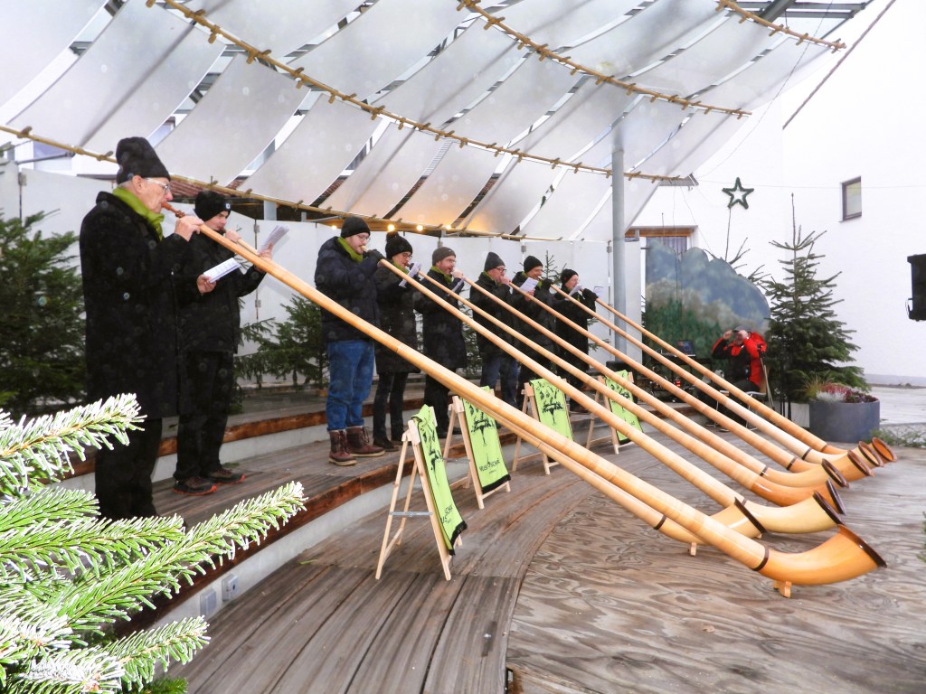 Alpenhornbläser, die bei einer Eröffnungsfeier auf einer Bühne spielen, umgeben von Weihnachtsbäumen.
