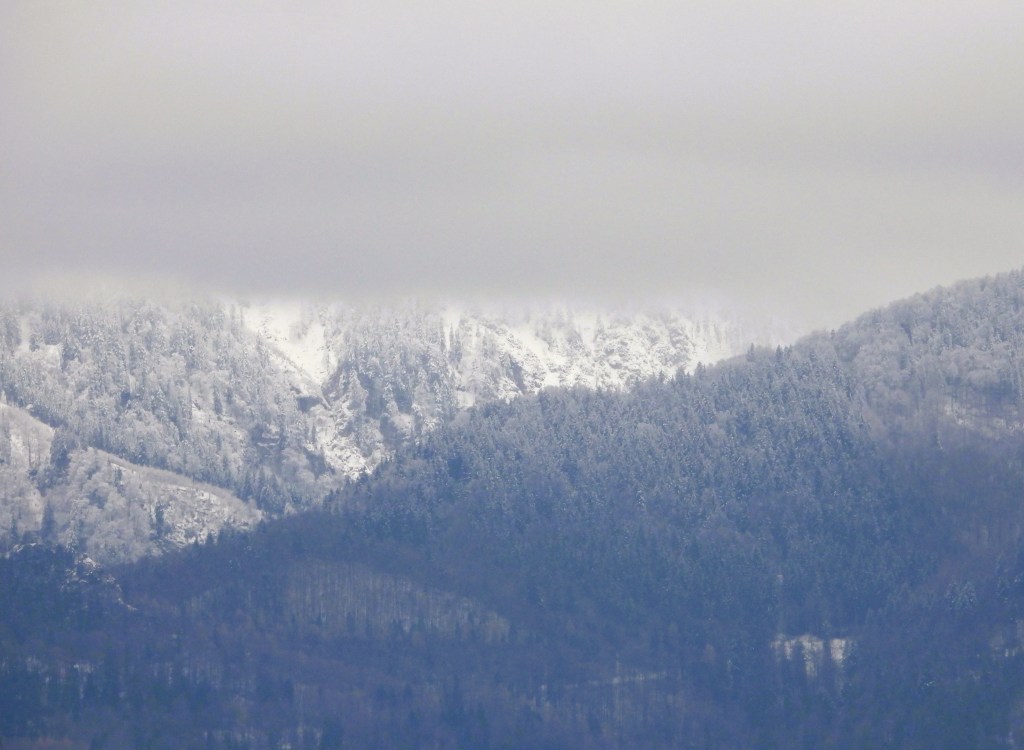 Schneebedeckte Berge unter einem grauen Wolkenhimmel, mit Nebelschwaden über den Gipfeln.