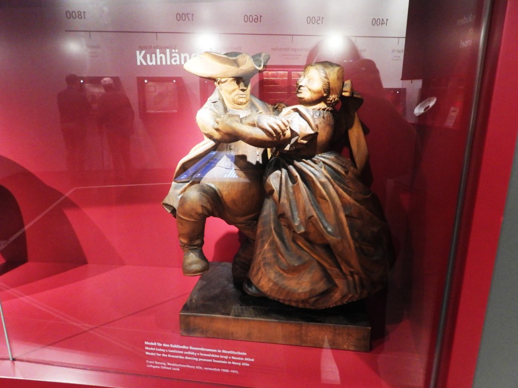 Holzskulptur eines tanzenden Bauernpaars in einem Ausstellungsraum im Sudetendeutschen Museum.