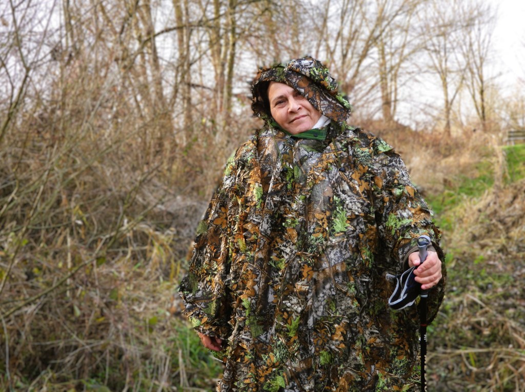 Eine Person steht in der Natur und trägt eine Tarnkleidung, um beim Beobachten von Vögeln unauffällig zu bleiben. Die Umgebung ist mit Bäumen und trockenem Gras umgeben.