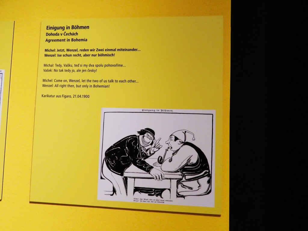 Ein Ausstellungstext aus dem Sudetendeutschen Museum mit einer Illustration, die eine Karikatur eines Gesprächs zwischen zwei Männern zeigt, umgeben von gelben Wänden.