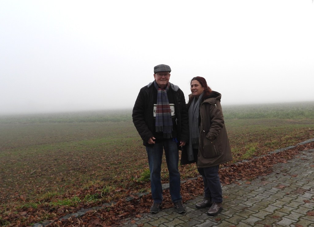 Ein Paar steht in einem nebligen Landschaftsbereich, umgeben von abgefallenen Blättern und einer grünen Wiese im Hintergrund. Beide tragen warme Kleidung und lächeln freundlich in die Kamera.
