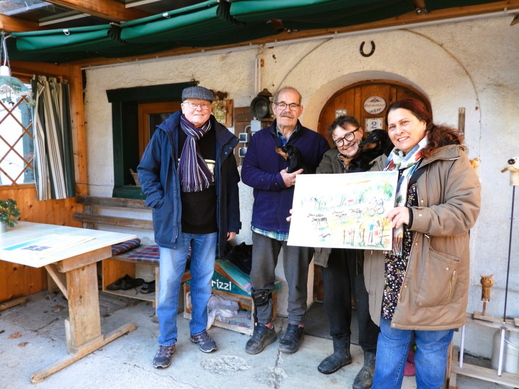 Eine Gruppe von vier Personen steht vor einem Holzgebäude. Zwei Männer und zwei Frauen halten einen Hund und ein gemaltes Bild in der Hand, während sie lächeln. Im Hintergrund sind Holzmöbel und eine dekorative Wand zu sehen.
