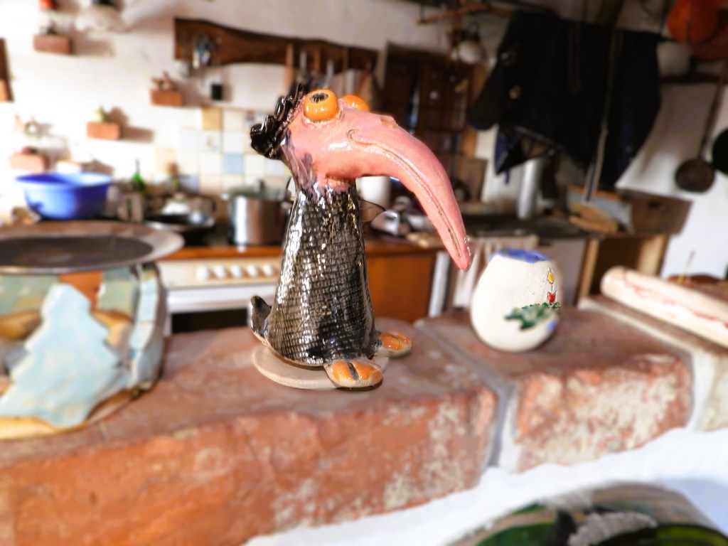 Eine bunte keramische Figur eines Vogels mit einem langen Schnabel und großen Augen, auf einer Steinplatte in einer rustikalen Umgebung platziert.