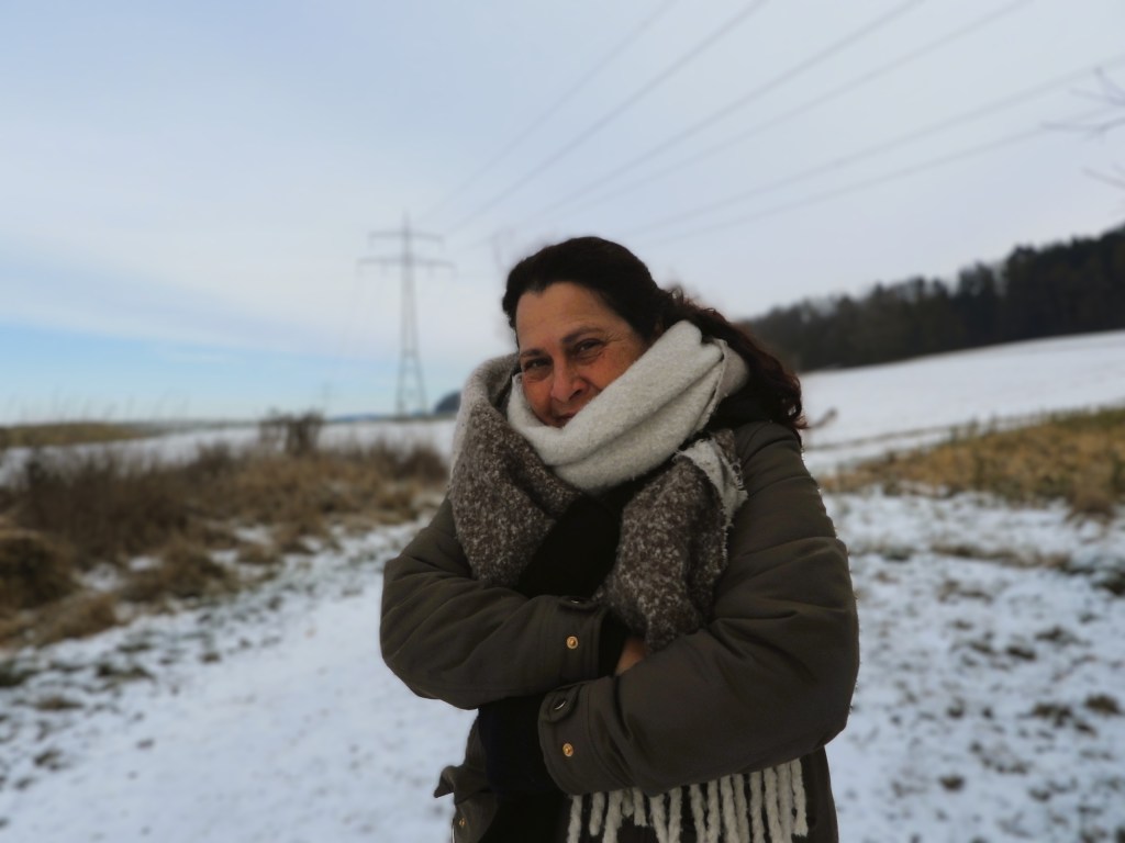 A woman standing outdoors in a snowy landscape, smiling and bundled up in a warm coat and scarf, with power lines and trees in the background.