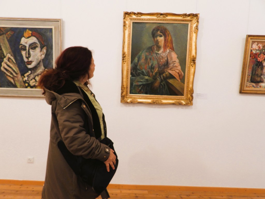 Eine Frau betrachtet ein Gemälde in einer Kunstgalerie, während sie eine Tasche trägt. Im Hintergrund sind weitere Gemälde an der Wand zu sehen.