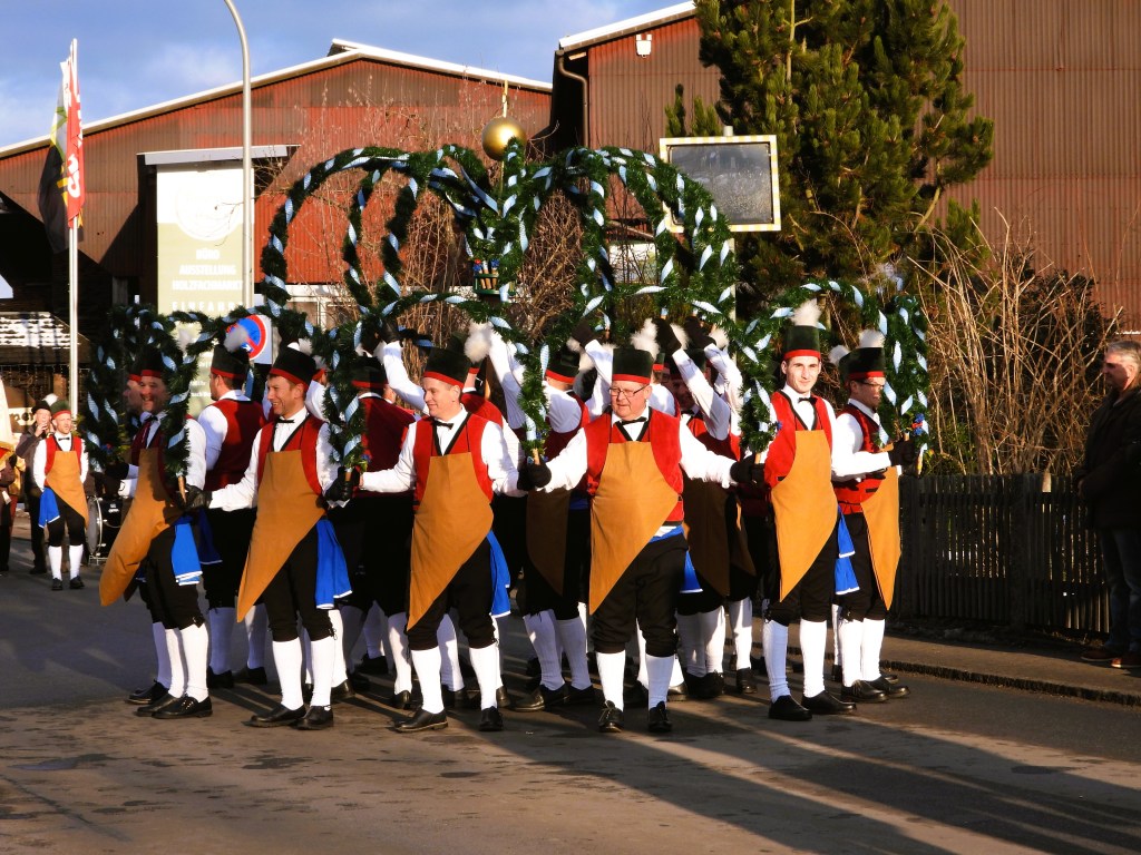 Eine Gruppe von festlich gekleideten Männern in traditioneller Tracht, die mit dekorierten Kränzen während eines Umzugs marschieren.