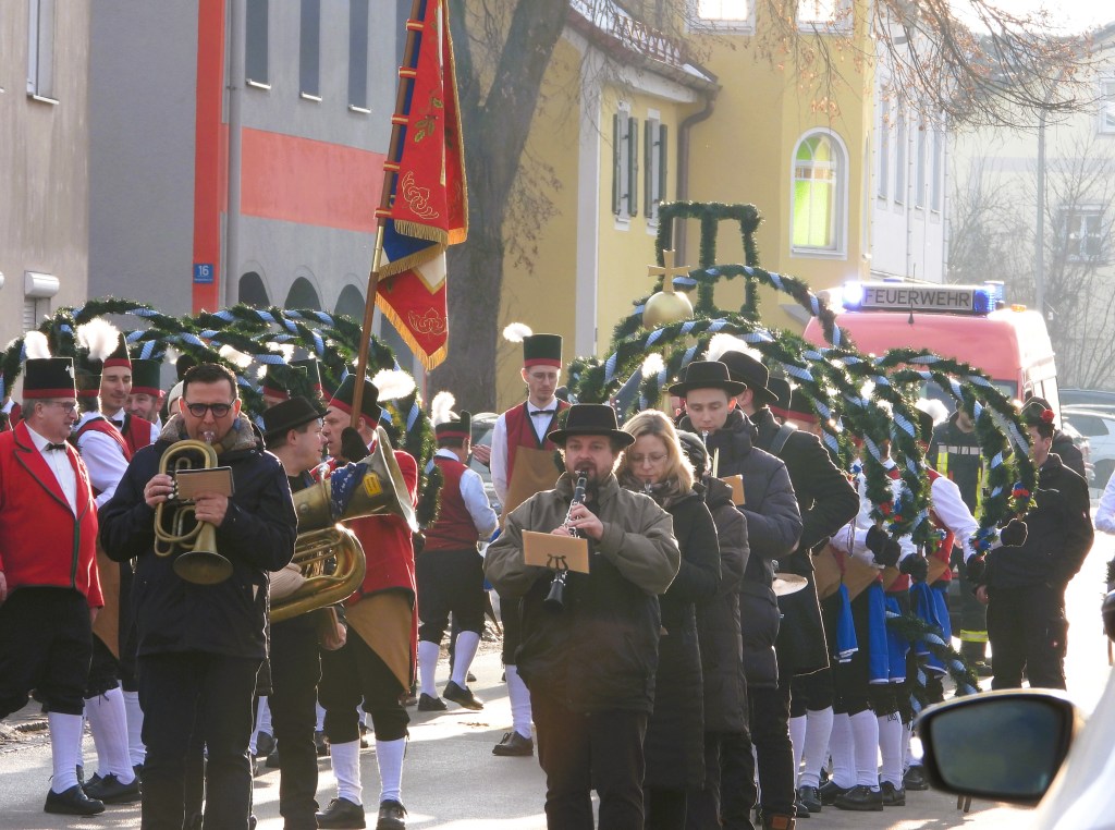 Eine Gruppe von Musikern und Feiernden in traditioneller Tracht, die während eines Festumzugs auf einer Straße marschieren. Eine Person spielt ein Blechblasinstrument, während andere Musikinstrumente in Händen gehalten werden. im Hintergrund ist ein Feuerwehrfahrzeug sichtbar.