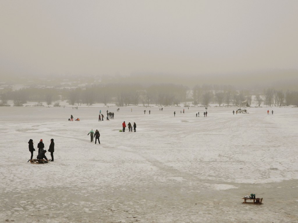 Ein winterlicher See mit schneebedecktem Eis, auf dem Menschen Schlittschuh laufen und Schlitten fahren. Die Landschaft ist neblig und grau, mit Bäumen und Häusern im Hintergrund.