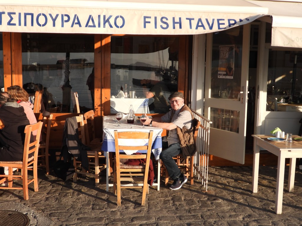 Ein älterer Mann sitzt an einem Tisch vor einer Fisch-Taverne mit einem Blick auf das Wasser. Er lächelt und hat ein Glas Wein vor sich.