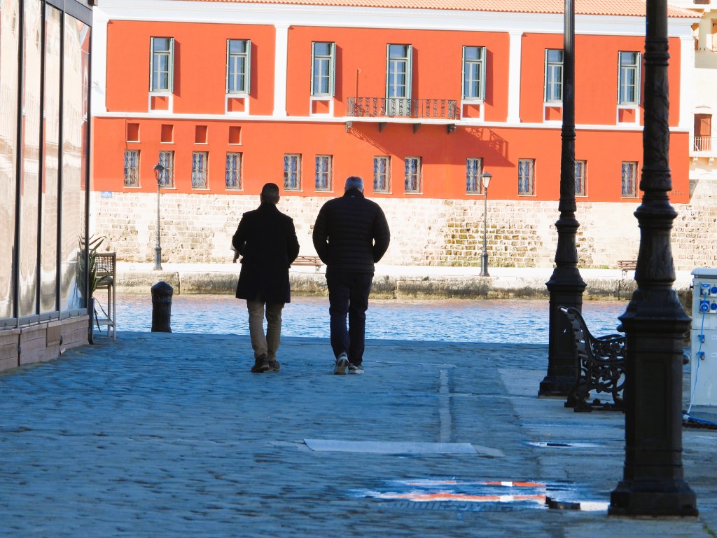 Zwei Männer gehen einen gepflasterten Weg entlang, vorbei an einem roten Gebäude mit Fenstern, am Wasser entlang.