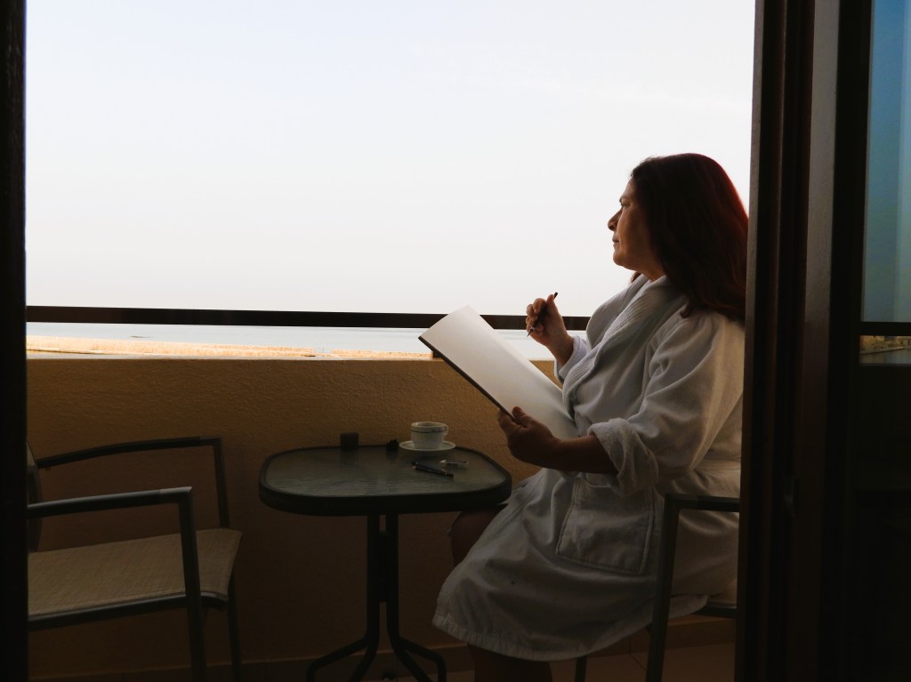 Frau in einem Bademantel sitzt auf einem Balkon und zeichnet in ein Skizzenbuch, während sie auf das Meer blickt.