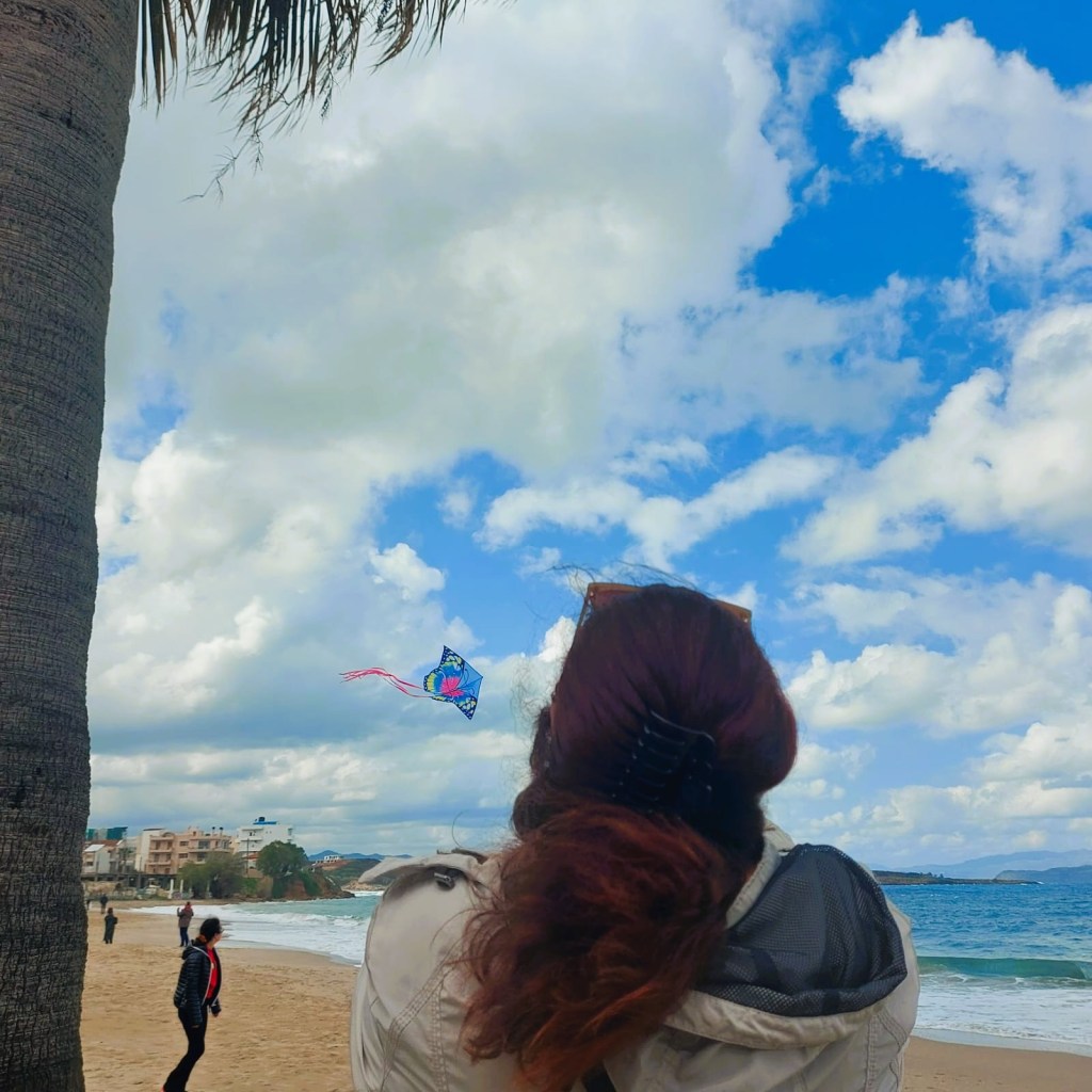 Eine Person steht am Strand und schaut einem bunten Drachen zu, der in einem blauen Himmel mit weißen Wolken fliegt.