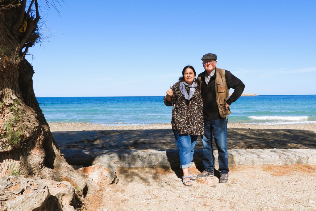 Ein Paar steht am Strand vor dem Meer, umgeben von einem klaren blauen Himmel und Bäumen.
