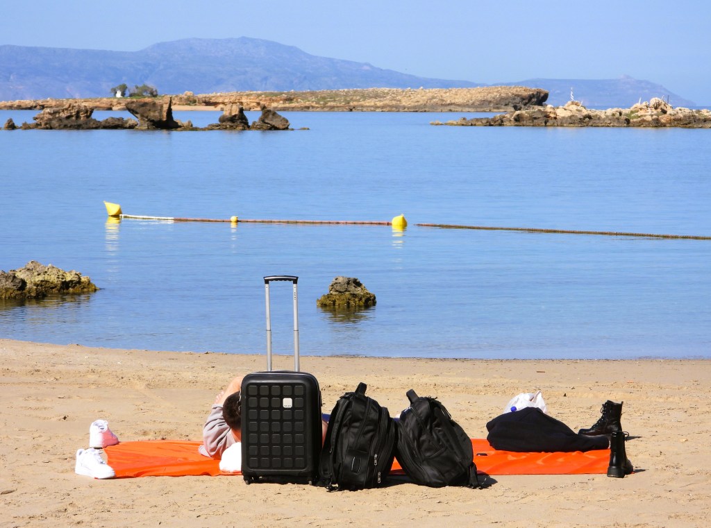 Personen liegen entspannt auf einem orangefarbenen Handtuch am Strand, während ein Koffer und zwei Rucksäcke in der Nähe stehen. Im Hintergrund ist ruhiges Wasser und Felsen zu sehen.