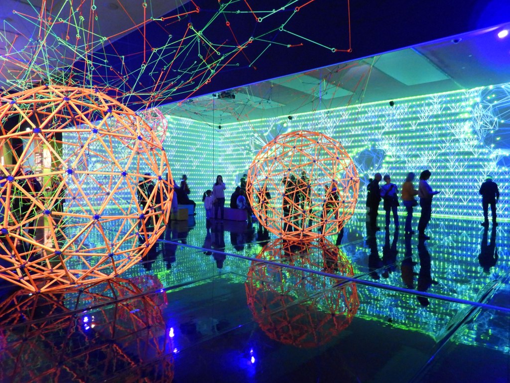 Innenansicht einer modernen Ausstellung mit geometrischen Skulpturen aus leuchtenden Materialien und einer farbenfrohen, visuell beeindruckenden Lichtinstallation im Hintergrund.