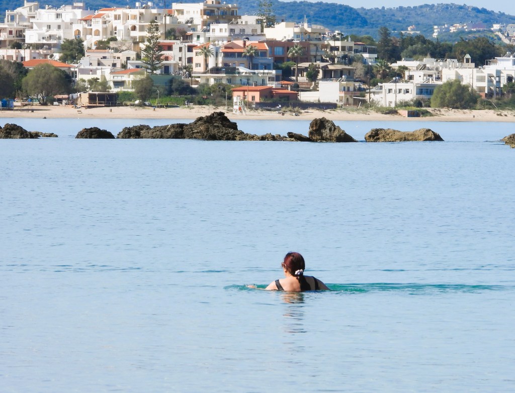 Eine Person schwimmt im klaren Wasser vor einer Küste mit Wohngebäuden im Hintergrund.