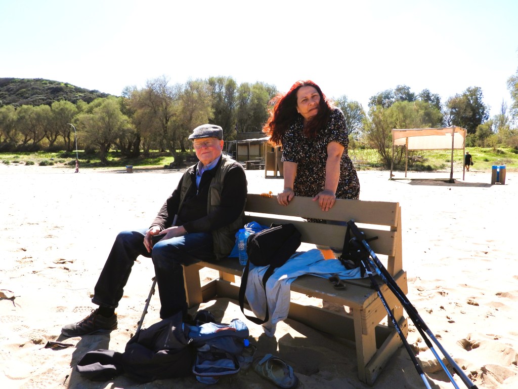 Ein Mann und eine Frau sitzen auf einer Bank am Strand. Der Mann trägt eine Mütze und eine Jacke, während die Frau ein blumiges Kleid trägt. Im Hintergrund sind Bäume und ein Stand mit einem Sonnenschutz zu sehen.