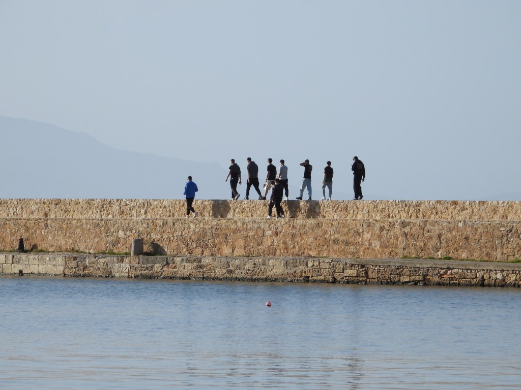 Menschen spazieren entlang einer Steinmauer am Wasser mit Bergen im Hintergrund.