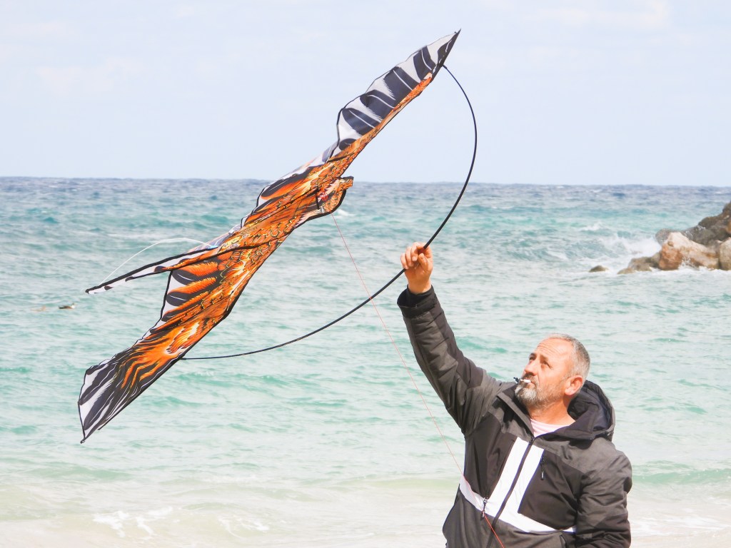 Ein Mann lässt einen bunten Drachen am Strand steigen, während das Meer im Hintergrund sichtbar ist.