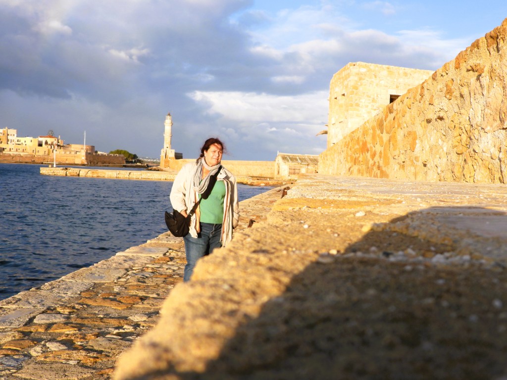 Eine Person geht entlang einer Steinmauer am Wasser, mit einem Leuchtturm im Hintergrund und einer stimmungsvollen Wolkenlandschaft.