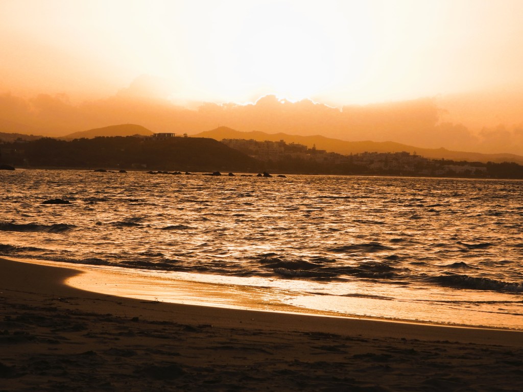 Sonnenuntergang am Strand mit Meer und Bergen im Hintergrund, warmes Licht und sanfte Wellen.