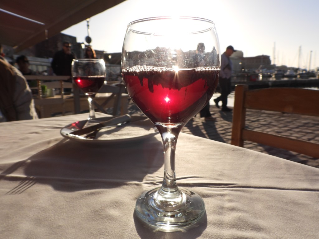 Ein Glas Rotwein auf einem Tisch mit Sonnenlicht, im Hintergrund sind Personen und ein Hafen zu sehen.