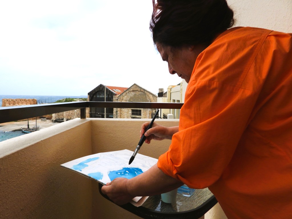 Eine Person in einem orangefarbenen Hemd malt auf einem Balkon mit Blick auf das Meer und historische Gebäude.